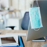 A Homedics Single Use Ear-Loop Face Mask hangs on the edge of a computer monitor screen in an office
