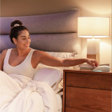 Dove | Homedics Portable Sound Machine with Night Light placed on a bedside table, while a woman relaxes in bed.