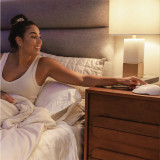 Cloud | Homedics Portable Sound Machine with Night Light placed on a bedside table, while a woman relaxes in bed.